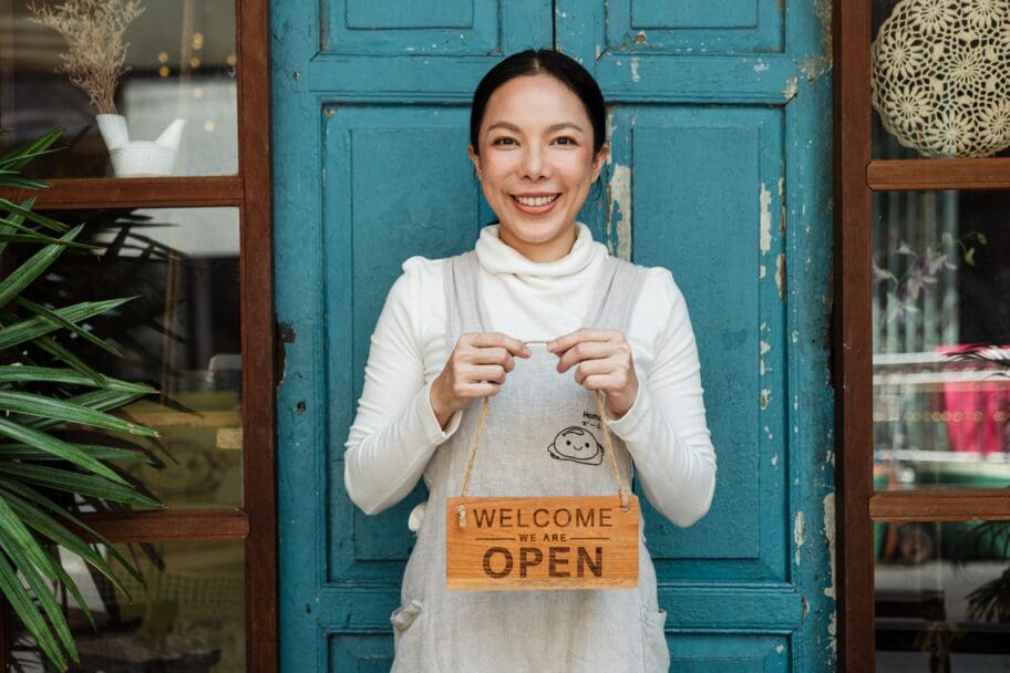 A smiling asian woman holding a welcome sign in front of a blue door, representing a small business.