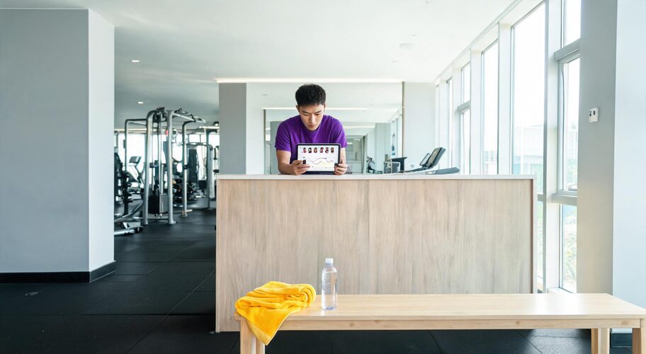 An empty early-morning gym floor with a single water bottle and towel left on a bench, while a coach by the reception counter checks AI for gym membership cancellations on a phone.
